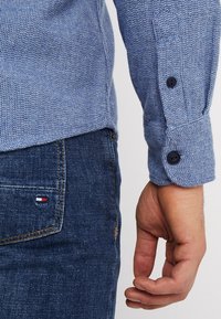 Camicia blu con texture e polsini con bottoni, indossata con jeans di denim scuro caratterizzati da un piccolo logo sulla tasca posteriore.