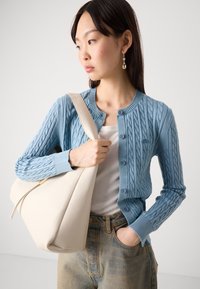 Light blue cable-knit cardigan with round neck and button closure. Model holds a large cream handbag, wearing a white top and denim jeans.