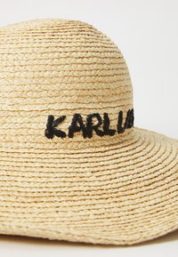 Širokobrim šešir od slame u prirodnoj boji s crnim vezom "KARL." Ima čvrsto tkanu teksturu i zaobljeni vrh.
