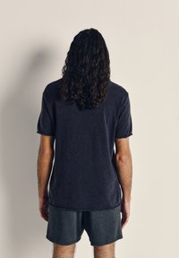 Person med långt lockigt hår, klädd i mörk kortärmad skjorta och shorts, står vänd mot en enfärgad ljus vägg.