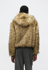 Casaco de pelo falso em tons castanhos e dourados com capuz, design texturizado e cropped. Cintura elástica preta; combinado com calças cinzentas às riscas.