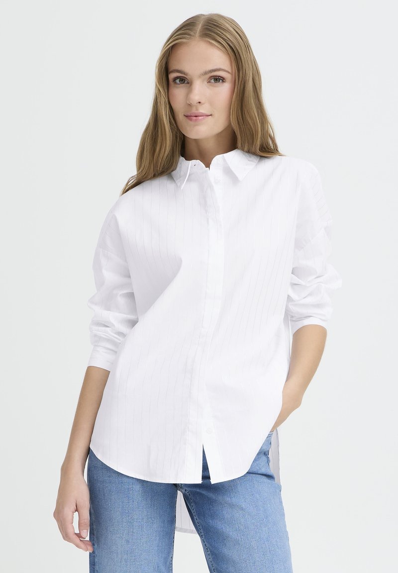 Valkoinen napillinen paita, jossa on pystysuorat raidoitukset, pitkät hihat ja kaareva helma. Yhdistetty vaaleansinihin farkkuihin. Silkkinen kankaan rakenne.