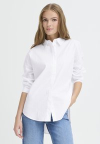 White button-up shirt with vertical pinstripes, long sleeves, and a curved hem. Paired with light blue jeans. Smooth fabric texture.