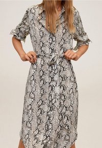 Femme nouant une ceinture sur une robe à boutons arrivant au genou, avec des manches retroussées et un motif en peau de serpent gris, noir et beige.