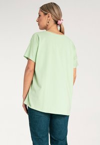 Jasnozielona bluzka krótki rękaw o luźnym kroju i zaokrąglonym dole, zestawiona z ciemnoniebieskimi dżinsami. Widok z tyłu podkreślający miękką teksturę materiału.