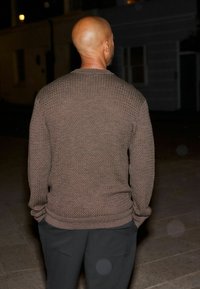 Brown knit sweater with a textured pattern and ribbed cuffs, paired with dark trousers, viewed from the back, in low light.