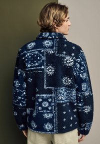 Fleece jack met een patchworkontwerp in marineblauw en witte bloemmotieven, voorzien van een kraag en een ontspannen pasvorm.