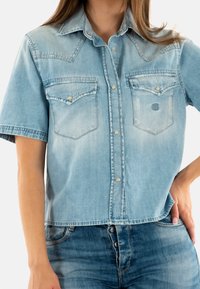 Camisa de mezclilla azul claro de manga corta con cuello puntiagudo, botones a presión y dos bolsillos en el pecho. Presenta un logo sutil en el bolsillo.