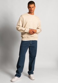 Felpa beige con logo impresso, colletto rotondo e polsini a coste, abbinata a jeans denim blu scuro e sneakers chiare.