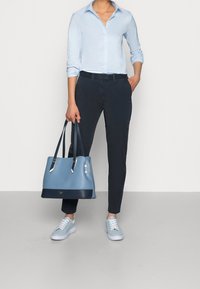 Persona vistiendo una camisa de botones azul claro, pantalones ajustados azul marino, zapatillas azul claro y sosteniendo un gran bolso azul claro y azul marino.