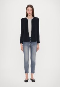 Zwart op maat gemaakt jasje met plooien, een ronde halslijn, gecombineerd met een lichtgrijze top en strakke blauwe jeans. Gladde textuur, eenvoudig ontwerp.