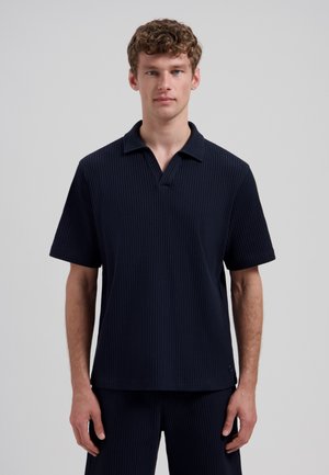 Jonge man met krullend haar, gekleed in een marineblauw, textuurgebreid poloshirt met korte mouwen en bijpassende short, staand tegen een effen achtergrond.