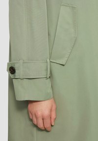 Lichtgroene trenchcoat met een manchet met een knoopdetail en een opgestikte zak. De stof heeft een gladde textuur en een gestructureerd ontwerp.