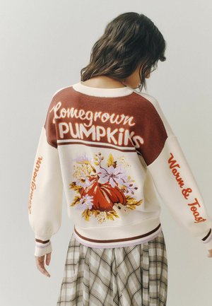 Woman wearing a cream and brown sweater with "Homegrown Pumpkins" text and pumpkin floral design on back, paired with a plaid skirt.