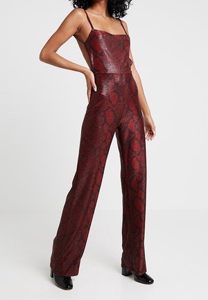 Femme portant une combinaison rouge sans manches à motif peau de serpent avec des jambes larges et des chaussures noires à talons, debout avec une main sur la hanche.