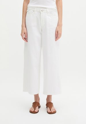 Vrouw draagt witte, wijde cropped broek en bruine sandaal met open teen, staand tegen een effen witte achtergrond.