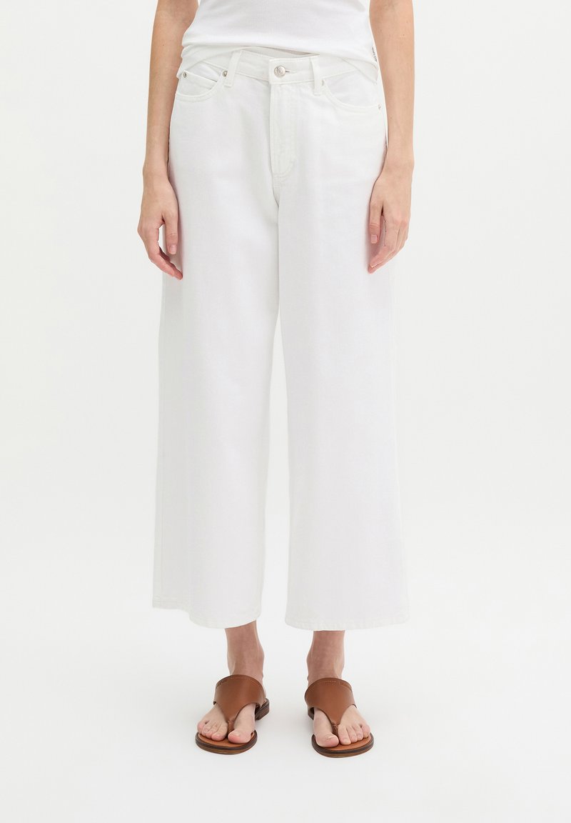 Woman wearing wide-leg white cropped pants and brown open-toe sandals standing against a plain white background.