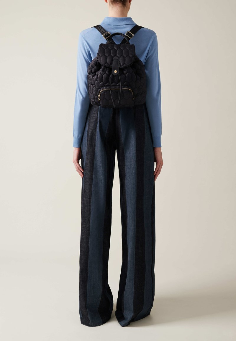 Black quilted backpack with a rounded top, gold zipper, and fabric straps. Paired with wide-leg trousers featuring vertical blue stripes.