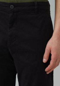 Schwarze Baumwollhosen mit glatter Textur, ausgestattet mit einem Knopfverschluss und seitlichen Taschen. Der Bund ist schlicht mit Gürtelschlaufen.