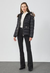 Jachetă puffer neagră cu fermoar frontal, bordură din blană artificială la glugă și buzunare laterale. Exteriorul texturat are detalii matlasate.