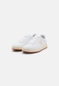 White leather sneakers with perforated toe, raised seams, and beige sole featuring cork texture; red text detail on side of left shoe.