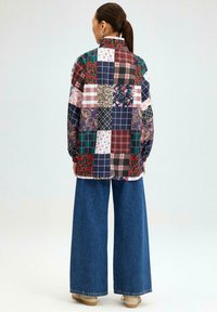 Multikleurige patchworkoverhemd met kraag; bevat bloemen- en ruitpatronen. Gepaard met denimbroek met wijde pijpen en beige schoeisel.