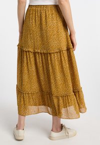 Gonna midi a strati in giallo senape con motivo floreale, orli arricciati e vita elasticizzata; abbinata a sneakers bianche.