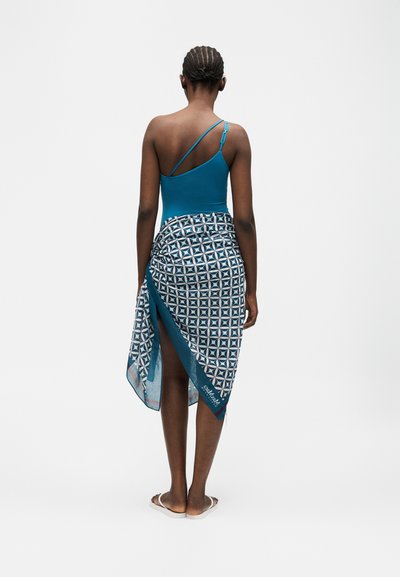 Γυναίκα με πλεξουδωτά μαλλιά που φοράει ένα τιρκουάζ τοπ με έναν ώμο και μια μπλε φούστα τύπου περιτύλιξης με γεωμετρικό σχέδιο, όρθια ξυπόλυτη σε λευκό φόντο.