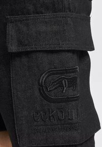 Zwarte denim shorts met een gestructureerde stof, opvallende patchzak en een reliëf logo ontwerp aan de linkerkant onderaan.