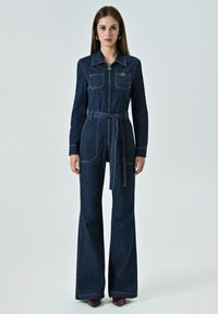 Dunkelblauer Denim-Overall mit langen Ärmeln, Reißverschluss vorne, zwei Brusttaschen, Taillenkordel und ausgestellten Beinen. Kontrastnähte sind vorhanden.