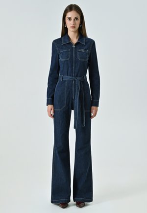 Combinaison en denim bleu foncé à manches longues, fermeture éclair sur le devant, deux poches poitrine, ceinture à la taille et jambes évasées. Détails de surpiqûres contrastantes.