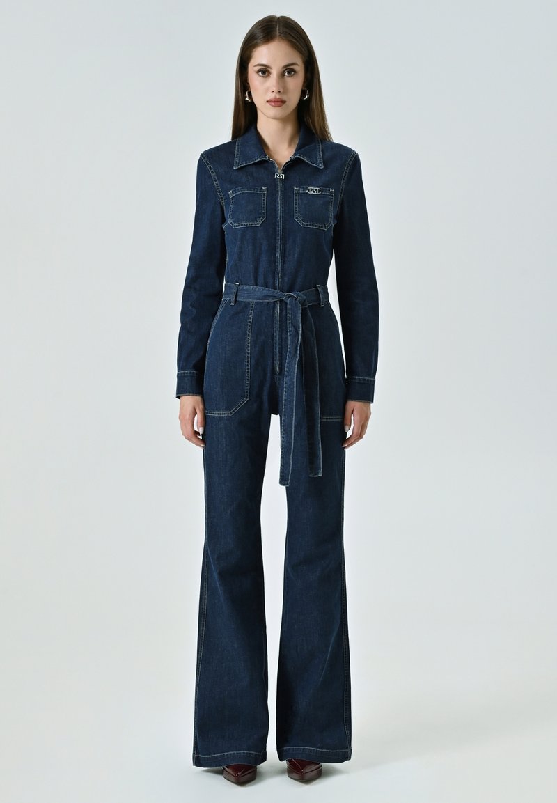 Dunkelblauer Denim-Overall mit langen Ärmeln, Reißverschluss vorne, zwei Brusttaschen, Taillenkordel und ausgestellten Beinen. Kontrastnähte sind vorhanden.