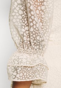 Blouse beige clair en tissu transparent, avec une texture à pois et des manches bouffantes élastiques. Les poignets froncés ajoutent du volume à la conception.