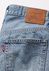 Šviesiai mėlynos džinsinės kelnės, turinčios rudą odinį etikėtę su reljefiniu tekstu ir mažą raudoną Levi's žymą ant užpakalinio kišenės.