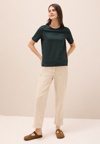 Camiseta de manga corta verde oscuro con texto en relieve, combinada con pantalones rectos de color beige claro y calzado marrón tipo slip-on, todo sobre un fondo neutro.