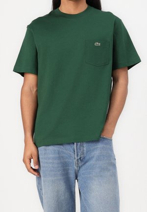 Mann trägt ein dunkelgrünes T-Shirt mit kurzen Ärmeln, Brusttasche und kleinem Krokodillogo, kombiniert mit hellblauen Jeans.