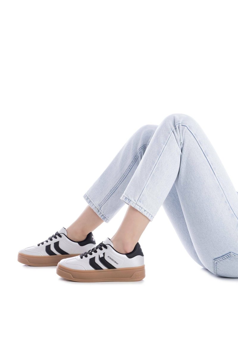 Zapatillas blancas y negras con suela de goma, que presentan rayas negras y parte superior texturizada. Combinadas con unos vaqueros recortados de color azul claro.