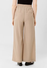 Béžové kalhoty širokého střihu vyrobené z měkké tkaniny s elastickým pasem a dvěma zadními kapsami. Lem končí těsně nad botami.