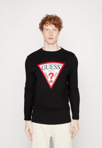 Zwarte langekoot T-shirt met een rood-witte driehoekige logo met de tekst "GUESS U.S.A." en accenten van "WASHED JEANS". Strakke pasvorm.