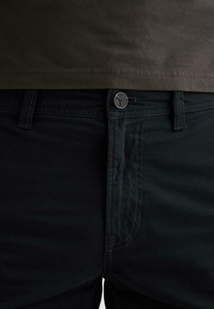 Detailný záber na čierne nohavice s gombíkom a pútkami na opasok, doplnené o tmavozelený olivový lem košele viditeľný nad pásom.