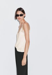 Kantafgewerkte, lichtbeige cami met dunne bandjes, gecombineerd met zwarte broek. Achtergrond is eenvoudig en lichtgrijs.