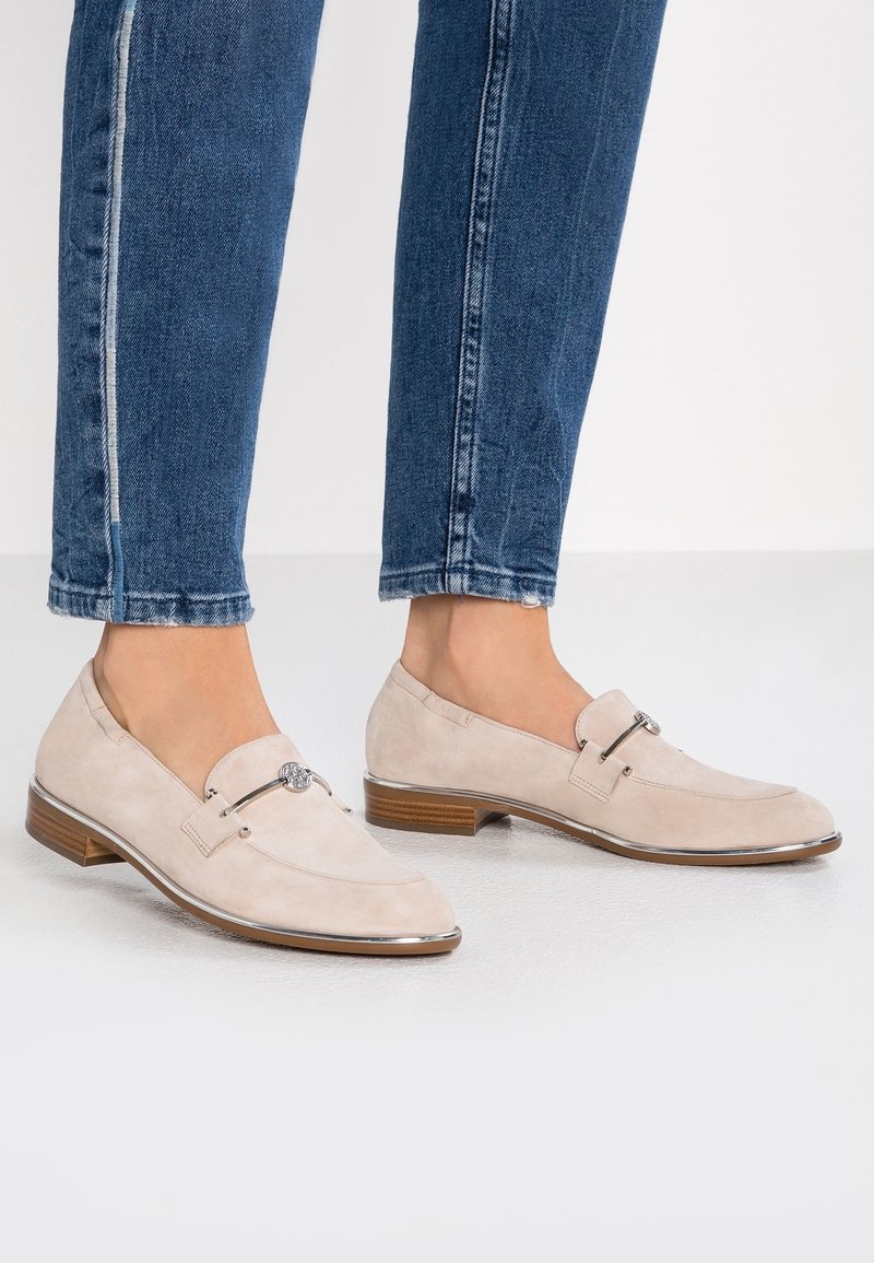 Mocassins en suède beige avec un bout arrondi et des détails en accent argenté. Talon bas empilé et garniture en cuir lisse. Portés avec un jean bleu.