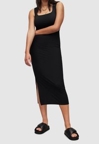 Robe midi noire ajustée avec un col carré, une fente latérale et une texture lisse; associée à des sandales noires épaisses à enfiler.