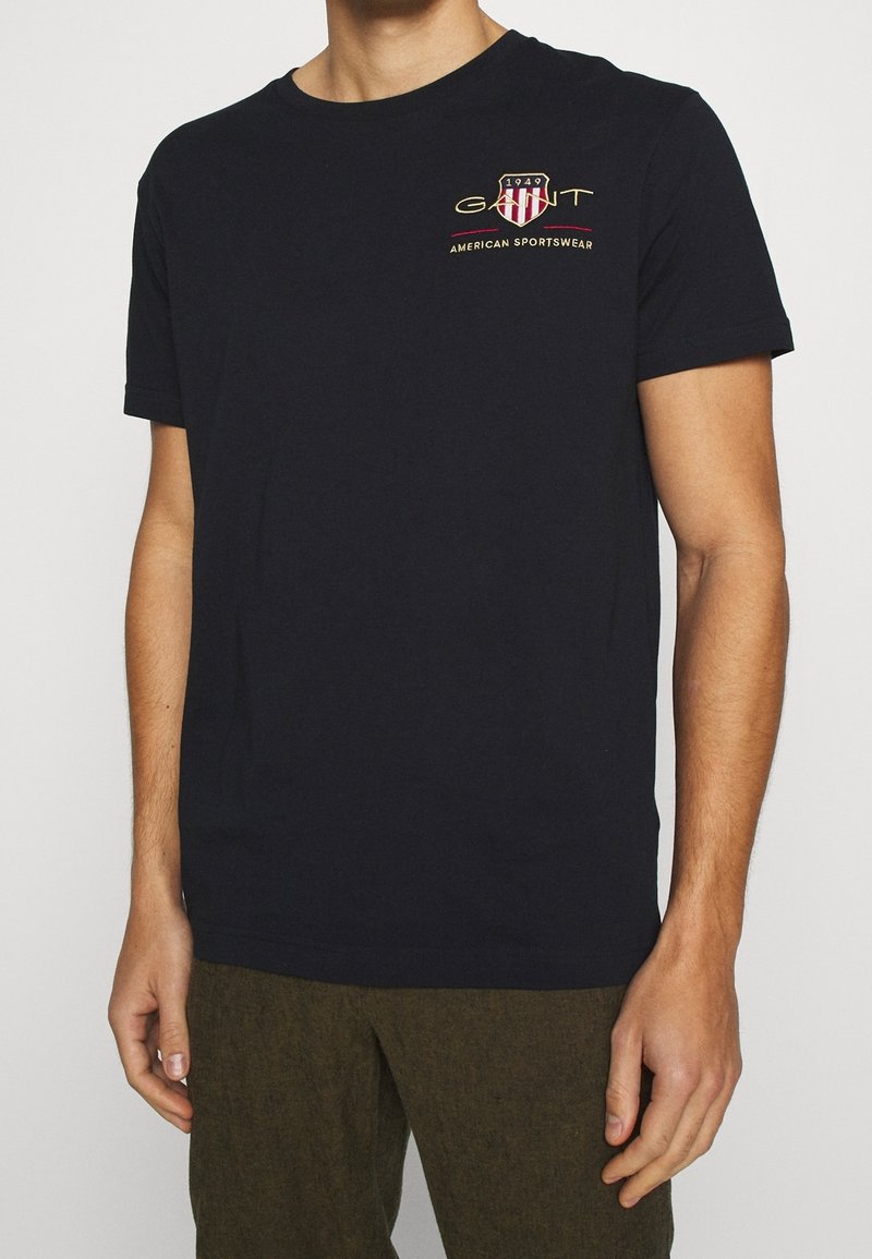 Homme portant un t-shirt noir à manches courtes avec un petit logo GANT rouge et or et du texte sur la partie supérieure gauche de la poitrine.