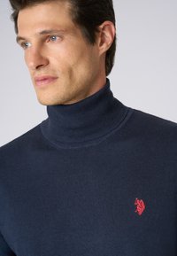 Maglione a dolcevita navy realizzato in morbido tessuto a maglia, con un logo ricamato rosso sul petto, superficie liscia e vestibilità aderente.