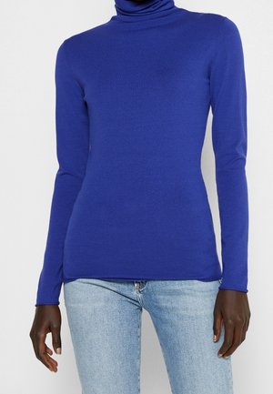 Persoon die een aangesloten koningsblauwe coltrui en lichtblauwe spijkerbroek draagt tegen een effen witte achtergrond.