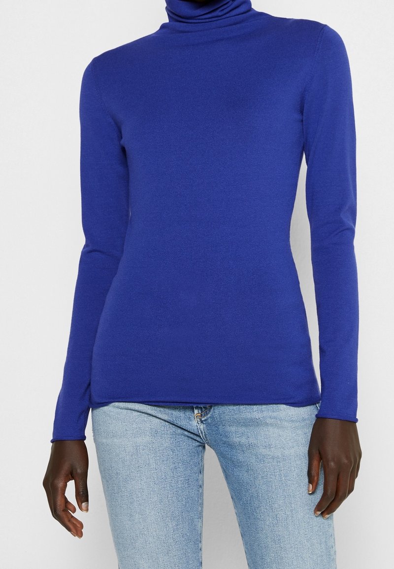 Personne portant un pull à col roulé ajusté bleu roi et un jean bleu clair, sur un fond blanc uni.