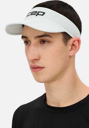 White sports visor with a wide brim and adjustable strap, featuring black "cep" logo. Model wears a plain black t-shirt.