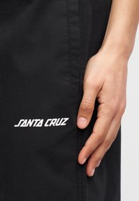 Χέρι που ακουμπά δίπλα σε μαύρο παντελόνι με λευκό λογότυπο "Santa Cruz" κοντά στην τσέπη στο πάνω μέρος του μηρού.