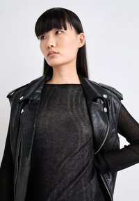 Gilet en cuir noir avec des accessoires en métal argenté superposé sur un haut sombre et texturé. La finition lisse du gilet contraste avec la qualité transparente du haut.